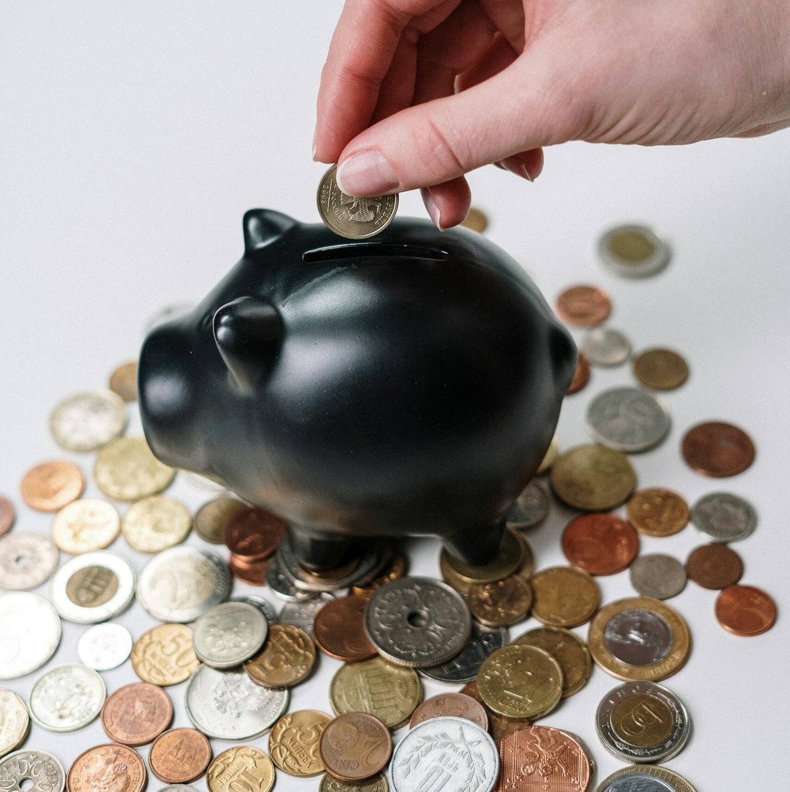 Hand inserting coin into a black piggy bank surrounded by various coins, symbolizing savings.